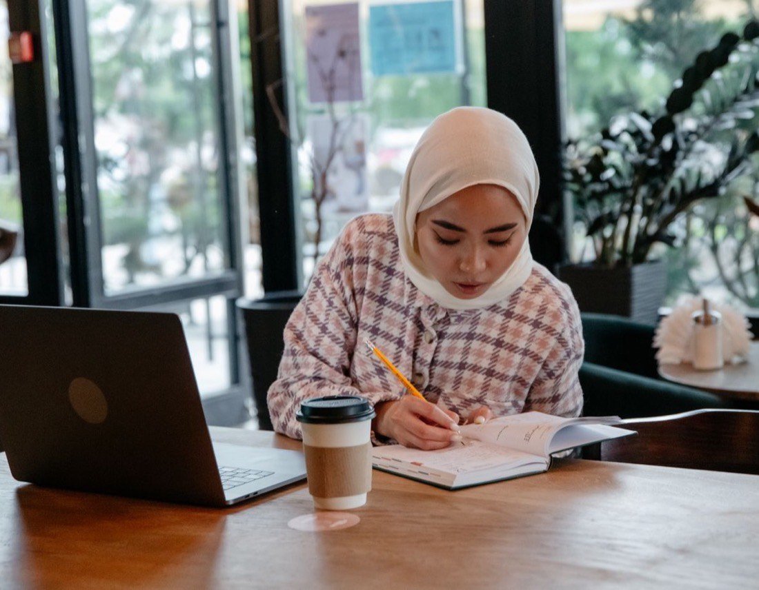 Peserta perempuan berhijab sedang mencatat sambil belajar di depan laptop